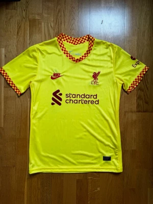 Gul Liverpool Nike matchtröja - Snygg gul Liverpool matchtröja från Nike med röda detaljer och rutigt mönster på krage och ärmslut. Tröjan har klubbmärke och Nike-logga på bröstet samt sponsortryck framtill. Tillverkad i lätt och ventilerande material, perfekt för fotboll eller träning.