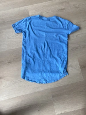Blå t-shirt med bröstficka - Säljer en enkel blå t-shirt i bomull med rund hals och korta ärmar. T-shirten har en liten bröstficka på vänster sida och en något längre, rundad nederkant baktill för en avslappnad look.
