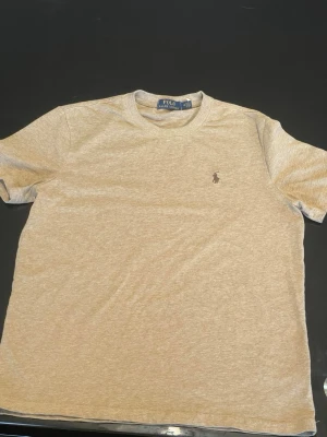 Beige t-shirt från Polo Ralph Lauren - Snygg beige t-shirt från Polo Ralph Lauren med klassisk rund hals och korta ärmar. T-shirten har det ikoniska broderade logotypen och är tillverkad i mjuk bomull för en skön känsla. Perfekt till jeans eller shorts. Passar S/M