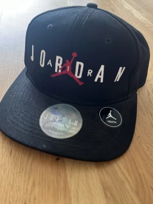 Air Jordan keps - Snygg svart keps från Jordan med den klassiska Jumpman-loggan i rött framtill och vit brodyrtext. Kepsen har en rak skärm. Märkt med 'Youth' på insidan och etikett. Perfekt för dig som gillar sportig streetstyle. Kepsen är justerbar och passar normalstort och mindre huvud. 