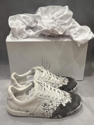 Maison Margiela vita sneakers med svart splash - Maison Margiela sneakers i vitt med unik svart splash-effekt på tå och sidor. Lågt skaft, klassisk siluett och snörning. Tillverkade i canvas och läder, med diskret logotyp på plösen. Perfekt för dig som gillar edgy och konstnärlig design.