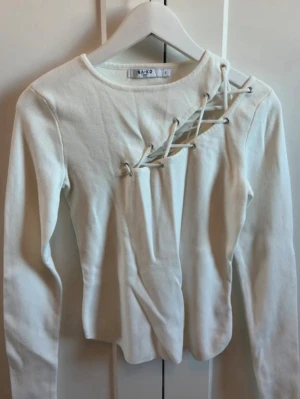 Vit långärmad topp  - Trendig vit långärmad topp från NA-KD med unik snörningsdetalj över bröstet. Toppen har rund halsringning och är tillverkad i ett mjukt bomullsmaterial. Perfekt för dig som vill sticka ut med en cool detalj.