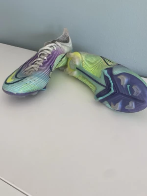Nike Mercurial färgglada fotbollsskor - Säljer ett par Nike Mercurial fotbollsskor med en riktigt snygg färgkombination i lila, turkos, gul och vitt. Skorna har en låg profil, snörning och tydliga dobbar för bra grepp på planen. Tillverkade i syntetmaterial för lätt känsla och maxad speed.