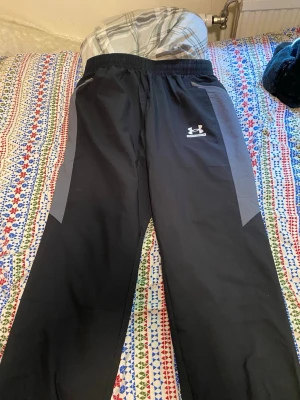 Svarta mjukisbyxor från Under Armour - Säljer ett par svarta mjukisbyxor från Under Armour med gråa detaljer längs sidorna. Byxorna har resår i midjan och logga på vänster lår. Perfekta för chill eller träning, tillverkade i ett mjukt och bekvämt material. PASSAR ÄVEM M