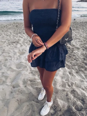 Meet me there dress - Säljer en marinblå axelbandslös kortklänning med smockad överdel och dubbla volanglager i kjolen. Kjolen har broderade detaljer och hålmönster som ger en cool och somrig vibe. Perfekt för dig som vill ha en unik och trendig look.