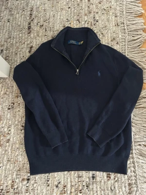 Mörkblå stickad tröja Polo Ralph Lauren - Mörkblå stickad tröja från Polo Ralph Lauren med halv dragkedja och klassisk broderad logga på bröstet. Tröjan har ribbade muddar och hög krage, perfekt för lager på lager. Materialet känns mjukt och passar till många olika stilar.