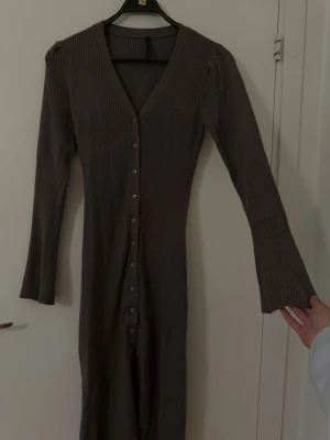 Brun ribbad långklänning med knappar - Snygg brun långklänning med ribbad struktur och v-ringning. Klänningen har långa ärmar och knappar hela vägen framtill, vilket ger en stilren och trendig look. Perfekt för dig som gillar en enkel men chic stil.