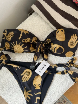 Helt oanvänd Svart och guld bikini från NA-KD - Snygg bikini från NA-KD i svart med guldiga abstrakta motiv. Bandeau-topp med knyt framtill och matchande bikinitrosa med knyt i sidorna. Tillverkad i mjukt syntetmaterial, perfekt för sommarens strandhäng.