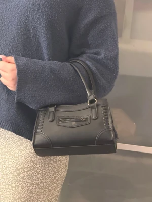 Svart handväska med dragkedjedetalj - Snygg svart handväska i konstläder med korta handtag och cool dragkedjedetalj framtill. Väskan har dekorativa sömmar och en stilren, boxig form som passar till många outfits. Perfekt storlek för det viktigaste och enkel att bära i handen eller på armen.