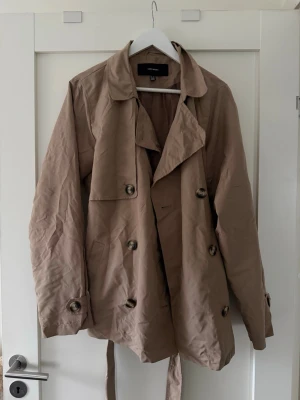 Beige trenchcoat från Vero Moda - Stilren beige trenchcoat från Vero Moda med klassisk dubbelknäppning, bred krage och bälte i midjan. Jackan har stora knappar och en avslappnad passform, perfekt för lager på lager. Tillverkad i ett lätt tyg som passar bra till vår och höst.