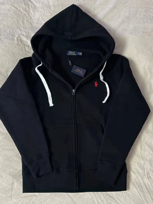 Svart hoodie från Polo Ralph Lauren - Svart hoodie med dragkedja från Polo Ralph Lauren. Klassisk design med vit dragsko i huvan och röd broderad logga på bröstet. Tillverkad i mjuk bomullsmix och har känguruficka framtill. Perfekt för en avslappnad och stilren look. Står storlek M men passar storlek S.