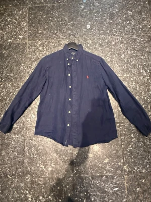 Mörkblå linneskjorta från Polo Ralph Lauren - Klassisk mörkblå skjorta från Polo Ralph Lauren i linne med button-down krage och lång ärm. Skjortan har knappar framtill och den ikoniska röda logotypen broderad på bröstet. Perfekt för en stilren och avslappnad look. Inte ink frakt