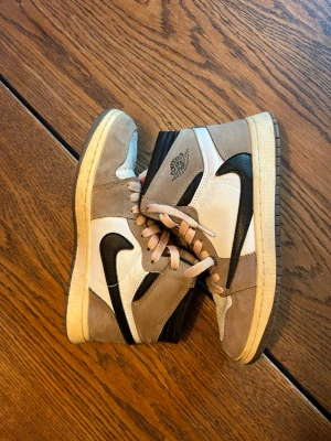 Nike Air Jordan 1 High cactus Jack - Säljer ett par Nike Air Jordan 1 med utanfvänd sula High sneakers i beige och vitt med svarta detaljer och swoosh. Skorna har klassisk hög siluett, perforerad tåbox och beige mockadetaljer. Tungan har Nike Air-logga i rött och skorna har ljusrosa snören. Perfekta för dig som gillar streetwear.