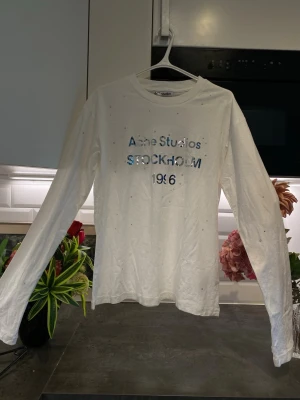 Vit långärmad topp Acne Studios - Vit långärmad topp från Acne Studios med tryck i silver och blått framtill där det står 'Acne Studios STOCKHOLM 1996'. Tunn bomullskvalitet och rund hals. Små glittriga detaljer över hela toppen för extra effekt.