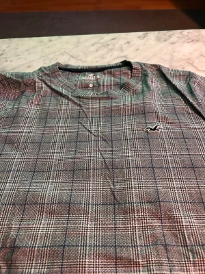  t-shirt Hollister - Snygg rutig t-shirt från Hollister med diskret logga på bröstet. T-shirten har korta ärmar och klassisk rund hals. Färgerna går i grått, blått och vitt, vilket ger en stilren och trendig look. Perfekt för dig som gillar mönstrade plagg med en modern touch.