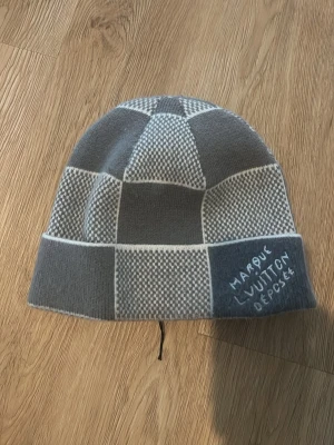 Gråvit rutmönstrad mössa - Säljer en stilren mössa i grått och vitt med rutmönster och broderad text på kanten. Mössan har en klassisk form och är tillverkad i mjukt stickat material som passar perfekt för kyliga dagar.