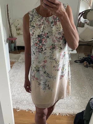 Blommig ärmlös kortklänning från Atmosphere - Söt ärmlös kortklänning med blommigt mönster i pastellfärger på en ljus, beige botten. Klänningen har rund halsringning och en rak, avslappnad passform. Perfekt för varma dagar och somriga tillfällen.