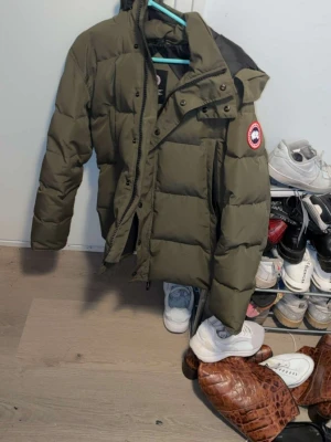 Olivgrön pufferjacka från Canada Goose - Säljer en olivgrön pufferjacka från Canada Goose med klassisk logga på ärmen. Jackan har hög krage, dragkedja och tryckknappar framtill samt stora, rejäla paneler för extra värme. Perfekt för kalla vinterdagar och riktigt snygg streetstil.