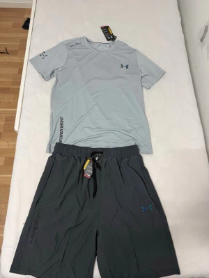 Under Armour träningsset t-shirt & shorts - Snyggt träningsset från Under Armour med ljusgrå t-shirt och mörkgrå shorts. T-shirten har diskret logga på bröstet och tryck på sidan, medan shortsen har elastisk midja med snörning och logga på benet. Perfekt för gymmet eller löprundan.