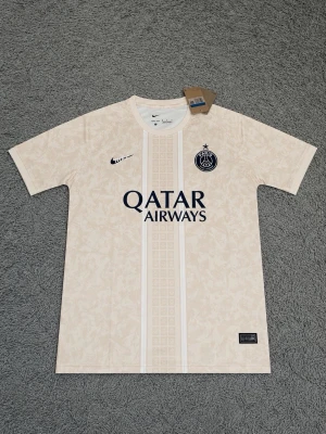 Beige PSG matchtröja från Nike - Snygg beige PSG matchtröja från Nike med diskret mönster, svart Nike-logga och PSG-emblem på bröstet. Qatar Airways tryckt i svart framtill. Perfekt för fotboll eller streetwear. Korta ärmar och rund hals.