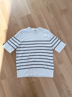 Vit randig topp med kort ärm - Vit topp med svarta horisontella ränder och korta ärmar. Klassisk rak passform och rund halsringning. Perfekt för en enkel och stilren look. Materialet känns mjukt och lätt, troligen bomull eller en bomullsblandning.