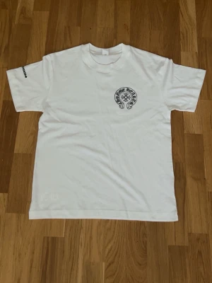 Vit Chrome Hearts t-shirt - Säljer en vit t-shirt från Chrome Hearts med svart tryck på bröstet och stor logga på ryggen. T-shirten har korta ärmar och rund hals. Materialet känns som mjuk bomull och det finns även ett litet tryck på ena ärmen.
