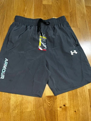  Under Armour shorts - Gråa reflektiva shorts från Under Armour med elastisk midja och snörning. Shortsen har reflex logga i regnbågsfärger och texten ARMOUR längs ena benet. Tillverkade i ett lätt och snabbtorkande syntetmaterial, perfekta för sport och träning.