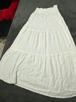 Vit långkjol med volanger - Säljer en vit långkjol med flera volanglager och smock i midjan. Kjolen har en luftig och flowy passform, perfekt för varma dagar. Enkel att matcha med olika toppar för en avslappnad stil.