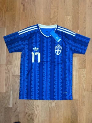 Blå Sverige fotbollströja Adidas - Säljer en blå fotbollströja från Adidas med Sveriges landslagsemblem, nummer 17 och namnet Gyökeres på ryggen. Tröjan har vita detaljer, klassiska Adidas-ränder på axlarna och ett unikt mönster. Perfekt för dig som vill stötta Sverige på match eller träning.