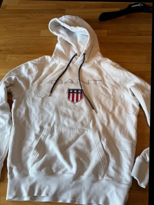 Vit hoodie från Gant med logga - Snygg vit hoodie från Gant med broderad logga och amerikanskinspirerat sköldmärke på bröstet. Hoodien har mörkblå dragsnören, stor känguruficka och mjukt bomullsmaterial. Perfekt för en avslappnad och trendig stil.