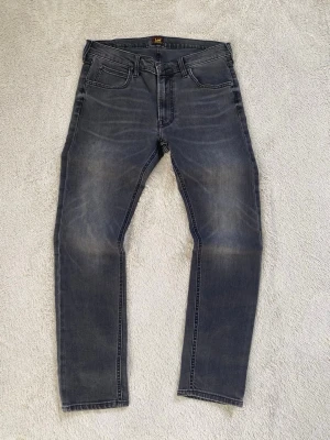 Grå slim fit jeans från Lee - ( W32 ) Snygga grå slim fit jeans från Lee med klassisk femficksmodell och diskreta slitningar på framsidan. Jeansen har normal midja, raka ben och Lee-logga på bakfickan. Perfekta för dig som gillar en stilren och avslappnad look.