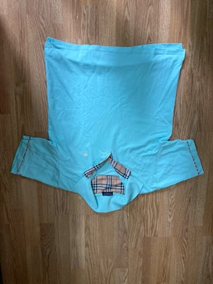 Ljusblå blus med rutiga detaljer - Trendig ljusblå blus från Burberry med volangdetaljer vid ärmarna och en unik rutig detalj framtill i klassiskt Burberry-mönster. Blusen har en lös passform och är perfekt för dig som gillar en avslappnad men ändå stilsäker look.