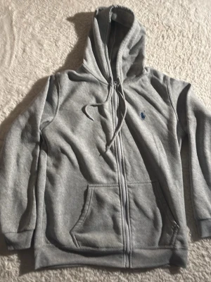 Grå zip hoodie från Polo Ralph Lauren - Snygg grå hoodie med dragkedja från Polo Ralph Lauren. Klassisk design med blå broderad logga på bröstet, huva med dragsko och stora fickor framtill. Tillverkad i mjuk bomullsblandning som är skön att ha på sig. Perfekt för en chill och avslappnad stil.
