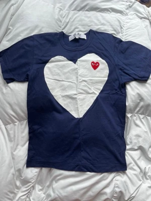 Blå Comme des Garçons Play t-shirt - Mörkblå t-shirt från Comme des Garçons Play med stort vitt hjärta på bröstet och ett mindre rött hjärta med ögon broderat ovanför. Klassisk rund hals och korta ärmar. Tillverkad i mjuk bomull för skön känsla.