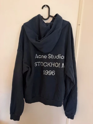  Acne Studios mörkblå hoodie - Säljer en mörkblå hoodie från Acne Studios med tryck på ryggen där det står 'Acne Studios STOCKHOLM 1996'. Hoodien har en stor huva och ribbade muddar vid ärmslut och nederkant. Perfekt för en avslappnad och cool stil.
