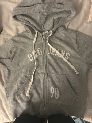 Grå zip-hoodie från Bug Jeans - Grå hoodie med dragkedja från Bug Jeans. Broderad text på bröstet och siffran 98 på ena fickan.  Skit snygg. Lx men passar mig som är S
