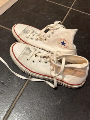Vita Converse Dam - Klassiska vita Converse Chuck Taylor All Star high-tops med röd rand längs sulan och blå logga på sidan. Skorna är i canvasmaterial med vita snören och gummisula. Perfekta för en avslappnad och cool stil.
