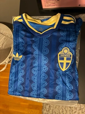 Blå Sverige fotbollströja Adidas - Säljer en blå fotbollströja från Adidas med Sveriges landslagsemblem på bröstet. Tröjan har gula detaljer vid kragen och axlarna samt ett retroinspirerat mönster i tyget. Perfekt för dig som vill stötta Sverige på match eller träning.