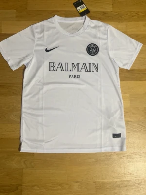 Vit PSG x Balmain t-shirt Nike - Snygg vit t-shirt från samarbetet mellan PSG och Balmain, med svart Nike-logga och PSG-emblem på bröstet. T-shirten har klassisk rund hals och korta ärmar. Materialet är mjukt och lätt polyester, perfekt för en sportig look.