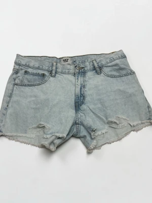 Ljusblå denimshorts med slitningar - Säljer ett par ljusblå denimshorts med råa kanter och slitna detaljer framtill. Klassisk femficksmodell med knapp och dragkedja. Perfekta för en avslappnad och trendig look.