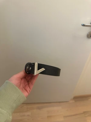 Svart Valentino bälte med V-spänne - Svart bälte från Valentino med ett stilrent V-format spänne i silverfärg. Passar någon som har storlek S/M.