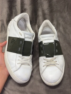 Valentino Opens, glansigt mörkgröna - Snygga vita sneakers från Valentino med ett brett, glansigt mörkgröna band över mitten. Skorna har klassisk låg profil, vita snören och diskret logga på plösen. Tillverkade i läder för en lyxig känsla och stilren look.