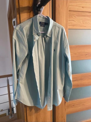 Ljusblå skjorta från Polo Ralph Lauren - Klassisk ljusblå skjorta från Polo Ralph Lauren med button-down krage och broderad logga på bröstet. Skjortan har lång ärm och knäppning framtill. Perfekt för en stilren och avslappnad look.
