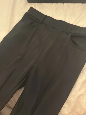 Svarta kostymbyxor med hög midja - Stilrena svarta kostymbyxor med hög midja och klassiska bälteshällor. Byxorna har raka ben och diskreta fickor framtill. Perfekta för en clean och dressad look. Materialet känns slätt och har en lätt glans.