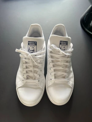 Adidas Stan Smith vita sneakers - Klassiska vita Adidas Stan Smith sneakers med blå detalj på hälen och perforerade ränder på sidorna. Skorna har lågt skaft, vita snören och är tillverkade i slätt läder. Perfekta för en clean och stilren look. Skorna är i princip helt nya och bara använda max 2 gånger. Skorna är använda och har någon skavnk (bilderna visar). Annars är de i helt nytt skick och kostar i original pris cirka 1000 kr, men jag säljer för 850 men priset kan diskuteras  