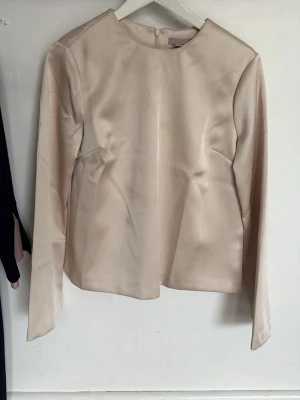 Beige satinblus från H&M - Stilren beige blus från H&M i glansigt satinmaterial. Blusen har rund halsringning, långa ärmar och dold dragkedja bak. Perfekt för en clean och elegant look. Materialet ger en lyxig känsla och passar till många olika outfits.