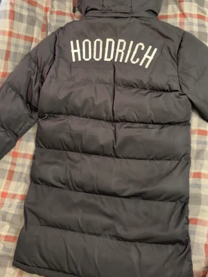 Svart lång dunjacka från Hoodrich - Säljer en svart lång dunjacka från Hoodrich med stor logga på ryggen och mindre logga på bröstet. Jackan har dragkedjor på fickorna, ribbade muddar vid ärmsluten och en huva. Perfekt för kalla dagar och riktigt snygg streetstil.