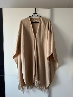 Beige ponchokappa med fransar - Stilren beige ponchokappa från C med lös passform och djup V-ringning. Kappan har en enkel knapp framtill och dekorativa fransar längs nederkanten. Tillverkad i mjukt tyg som ger ett snyggt fall. Perfekt för lager på lager och en avslappnad look.