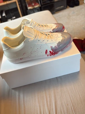 Vita sneakers med röd färgdetalj - Unika vita sneakers med grå och röd färgdetalj framtill som ger en cool splash-effekt. Skorna har klassisk låg silhuett, vita skosnören och en stilren design. Perfekta för dig som vill sticka ut med din stil