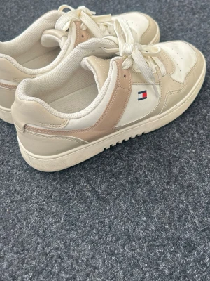 Beige Tommy Hilfiger sneakers - Snygga sneakers från Tommy Hilfiger i beige och vitt med klassisk logga på sidan. Skorna har lågt skaft, vita snören och ovandel i en mix av läder och textil. Perfekta för dig som gillar stilrena och tidlösa sneakers.